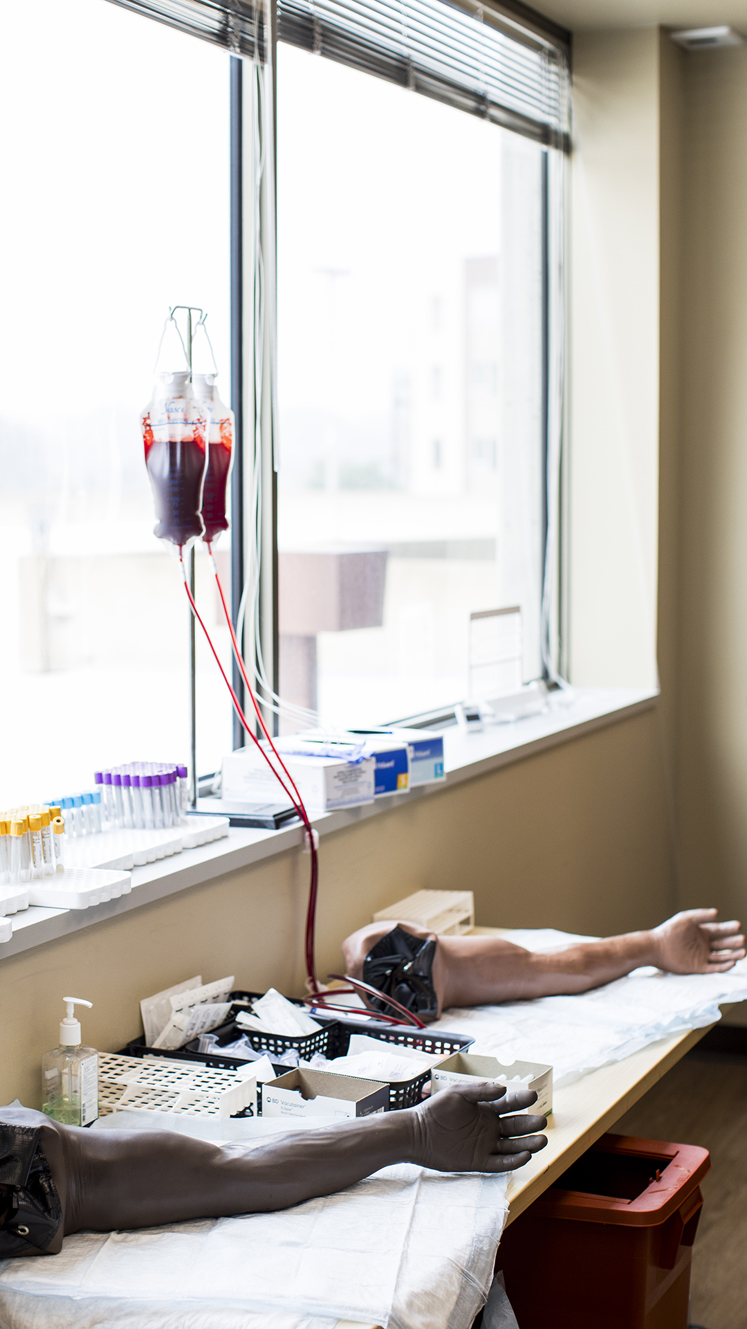 Two mannequin arms sit on top of absorbent pads on a table with a bottle of hand sanitizer and plastic trays filled with bandages in between them, with two IV bags hanging on a stand with tubes running down to one arm standing on a windowsill next to trays filled with plastic vials and in front of open blinds.