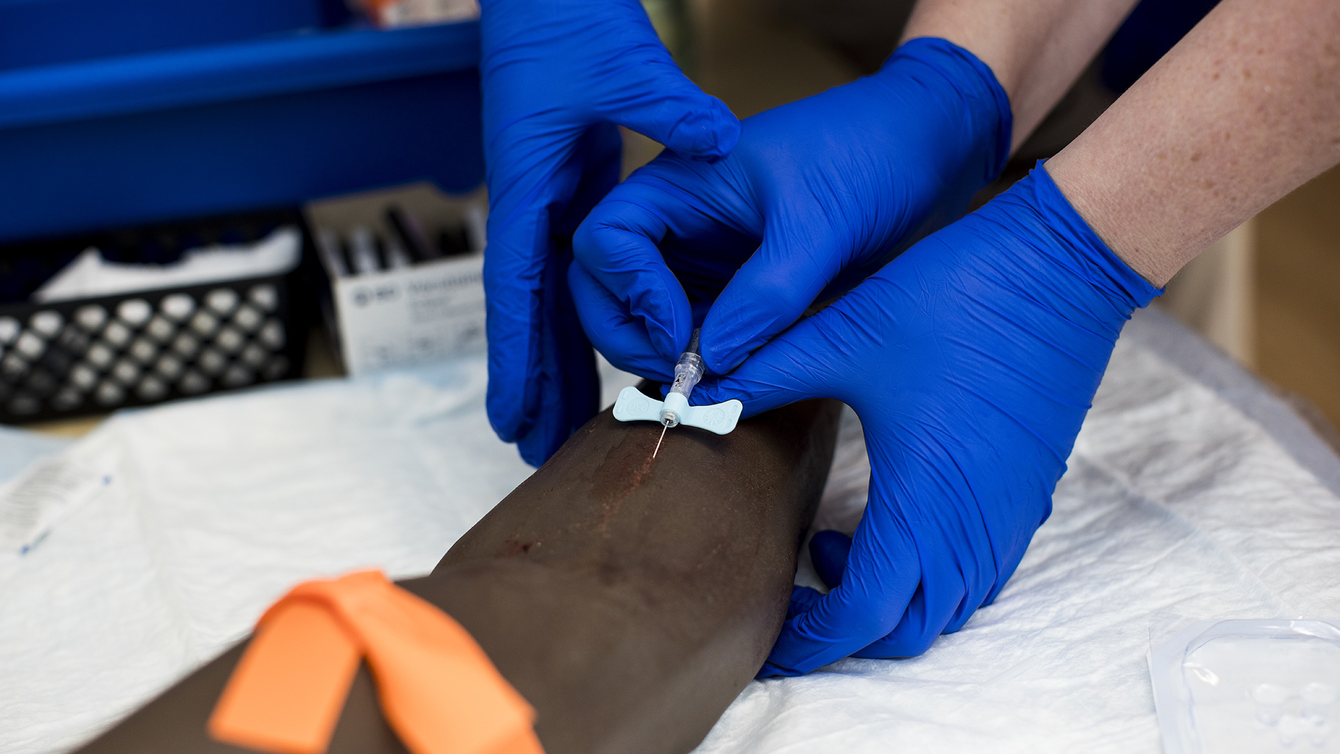 Two hands wearing sterile gloves insert a syringe into the forearm of a mannequin's arm sitting on an absorbent pad, with another hand wearing a sterile glove helping guide the action.
