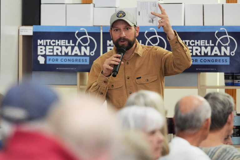 A man in a baseball hat and a tan shirt speaks to a crowd.