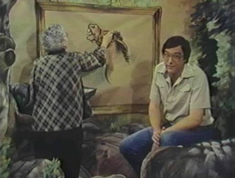 Screenshot from a 1970s tv show. A woman on the left is drawing a picture on a large backdrop while a man on the right talks to the camera.