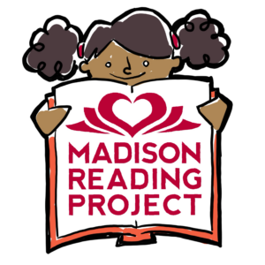 Little girl smiling as she peeks over the top of an open book decorated with the Madison Reading Project logo — a red heart nestled between open pages, with "MADISON READING PROJECT" beneath it in red letters. The girl has dark hair styled in two curly puff buns, and she holds the book open with both hands, orange showing on the edges of the pages.