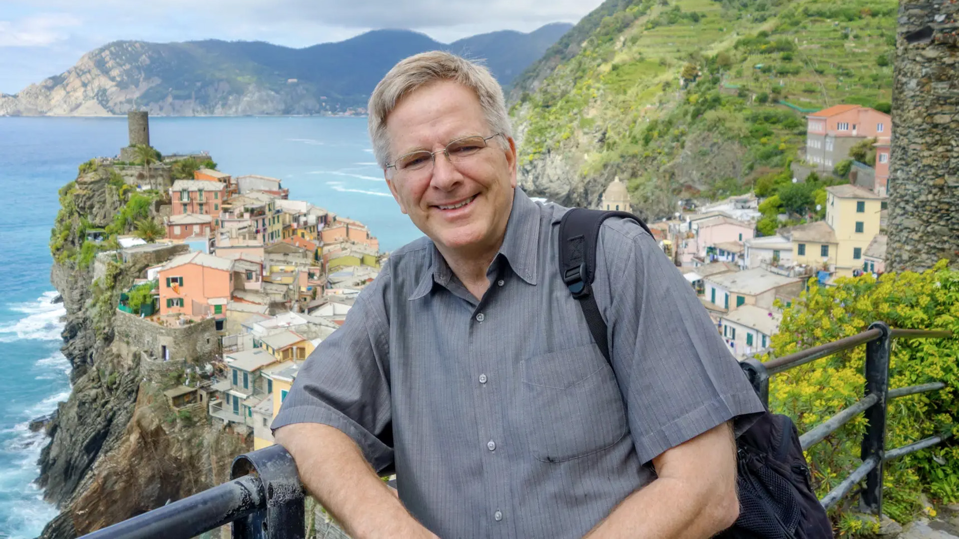 Rick Steves smiles at the camera, leaning on a railing overlooking Vernazza, a cliffside village in Italy's Cinque Terre. Colorful buildings cling to the rocky coastline above the sea, with green terraced hills rising in the background.