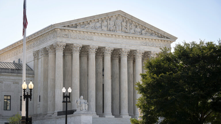 A U.S. flag is displayed on a flagpole in front of a stone masonry building with steps leading up to an entrance framed by a portico of columns upholding a pediment with sculptures of figures, with two painted metal lamp posts with two globes and a tree in the foreground.