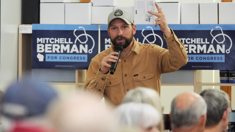 Mitchell Berman gestures with his left hand while speaking into a wireless microphone held in his right hand, while standing in front of multiple campaign signs with the words Mitchell Berman for Congress and a graphic of a stethoscope, with out-of-focus backs of heads of people facing him in the foreground.
