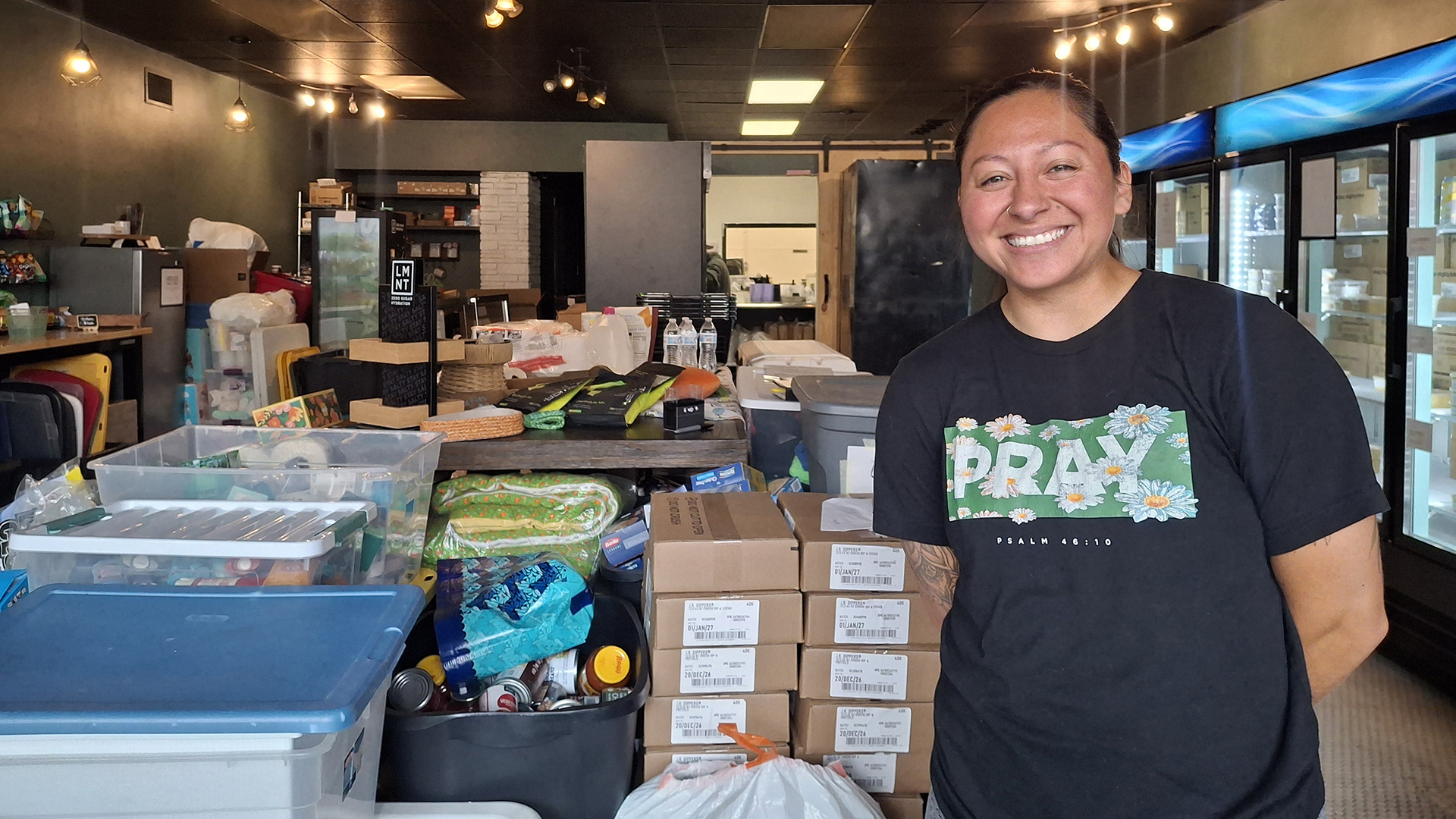 Destiny Atuna poses for a portrait while standing in front of stacks of items in bags, boxes and bins, with additional items stacked on a table in the background alongside additional storage bins, in a room with one wall lined with commercial glass door display refrigerators, more items stacked on the opposite wall, and track lighting and fluorescent lights in the ceiling.