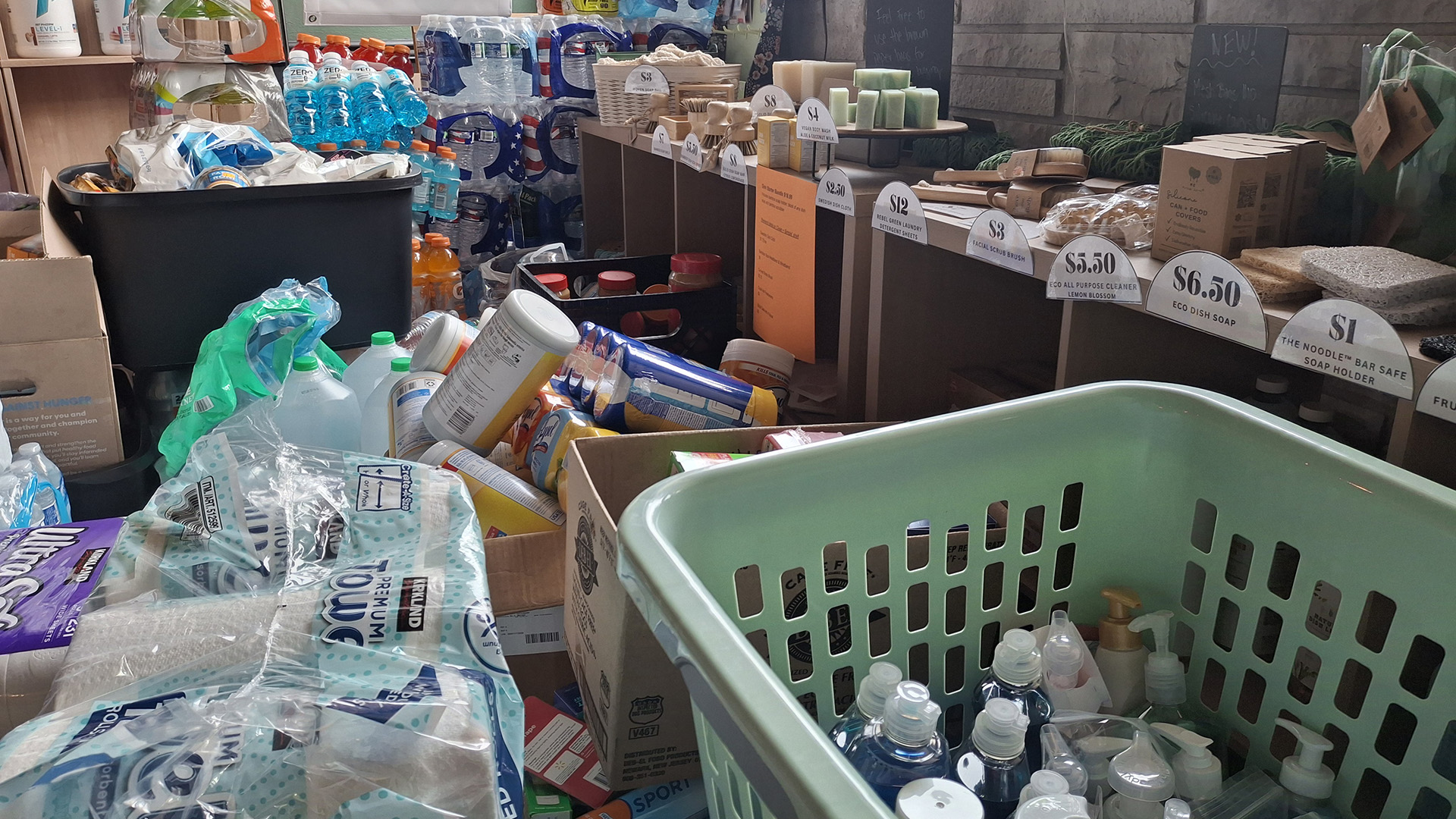 Different personal and home consumer proejcts, including paper towels, soaps, wipes, bottled water and electrolyte beverages sit in stacks in a room next to a row of shelves marked with price labels and stocked with different items.