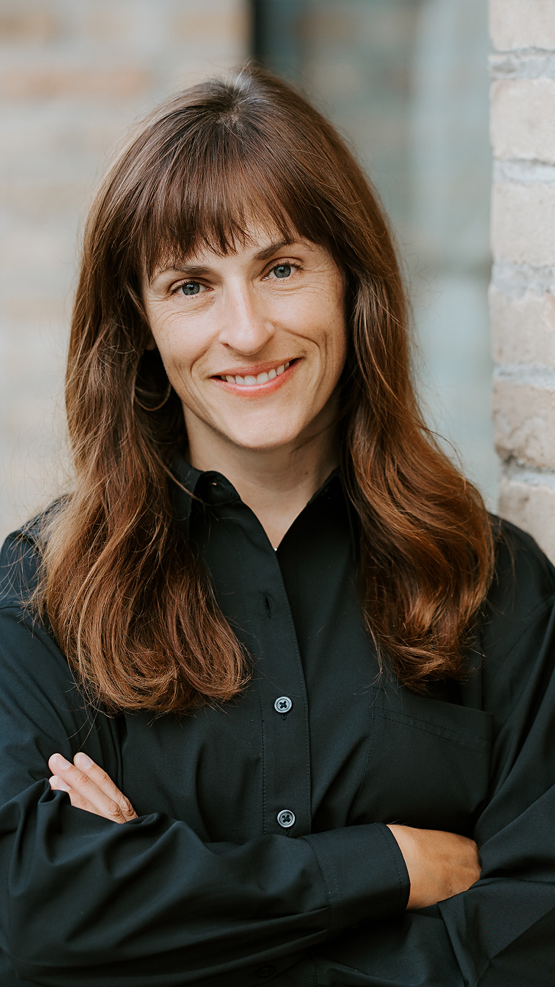Lora Glasel poses for a portrait while standing in front of a brick wall and window.