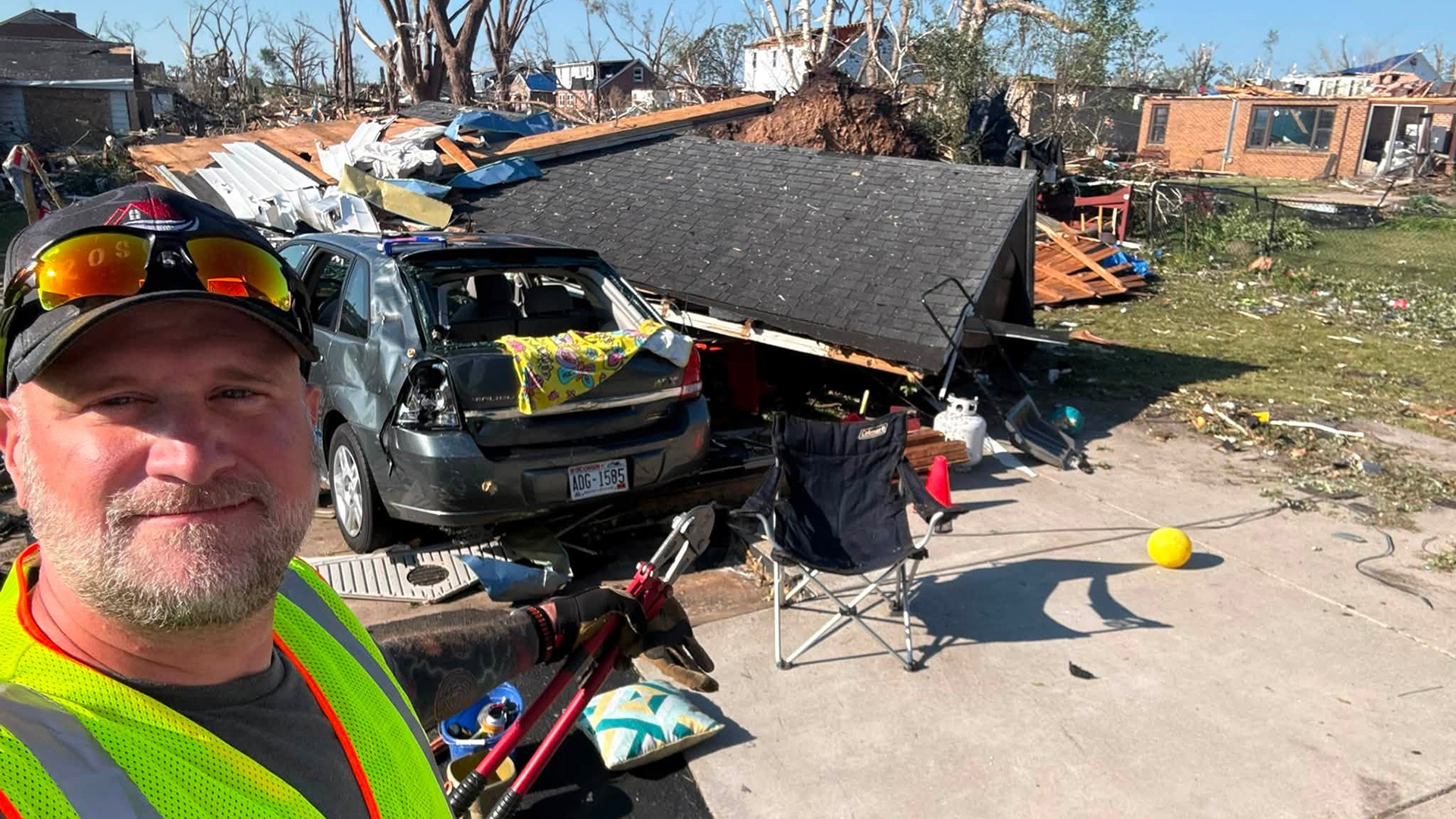 Shane Rosenow poses for a selfie while standing in front of a driveway with a garage roof and other debris covering and sitting next to a damaged car with broken windows, with more damaged structures in the background.