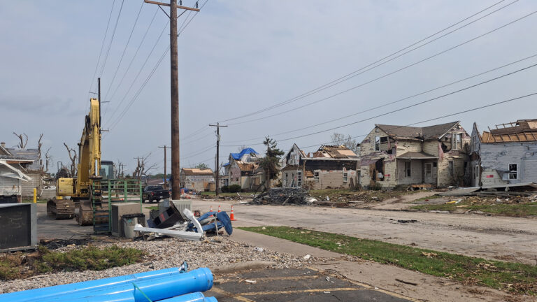 An excavator sits in a driveway next to a stack of debris on landscape stones and a sidewalk along a road lined with two parallel wood power line poles, with multiple houses with damaged siding, roofs and windows across the street, with tree trunks shorn of branches and leaves in the background and a stack of sewer pipes on a driveway in the foreground.