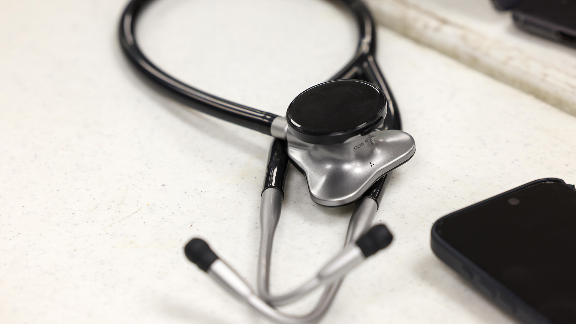 A stethoscope sits next to a smart phone on the surface of a table.
