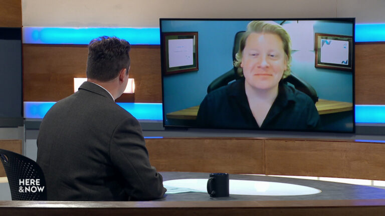 Zac Schultz sits at a desk on the Here & Now set and faces a video monitor showing an image of Andrew Mercil.