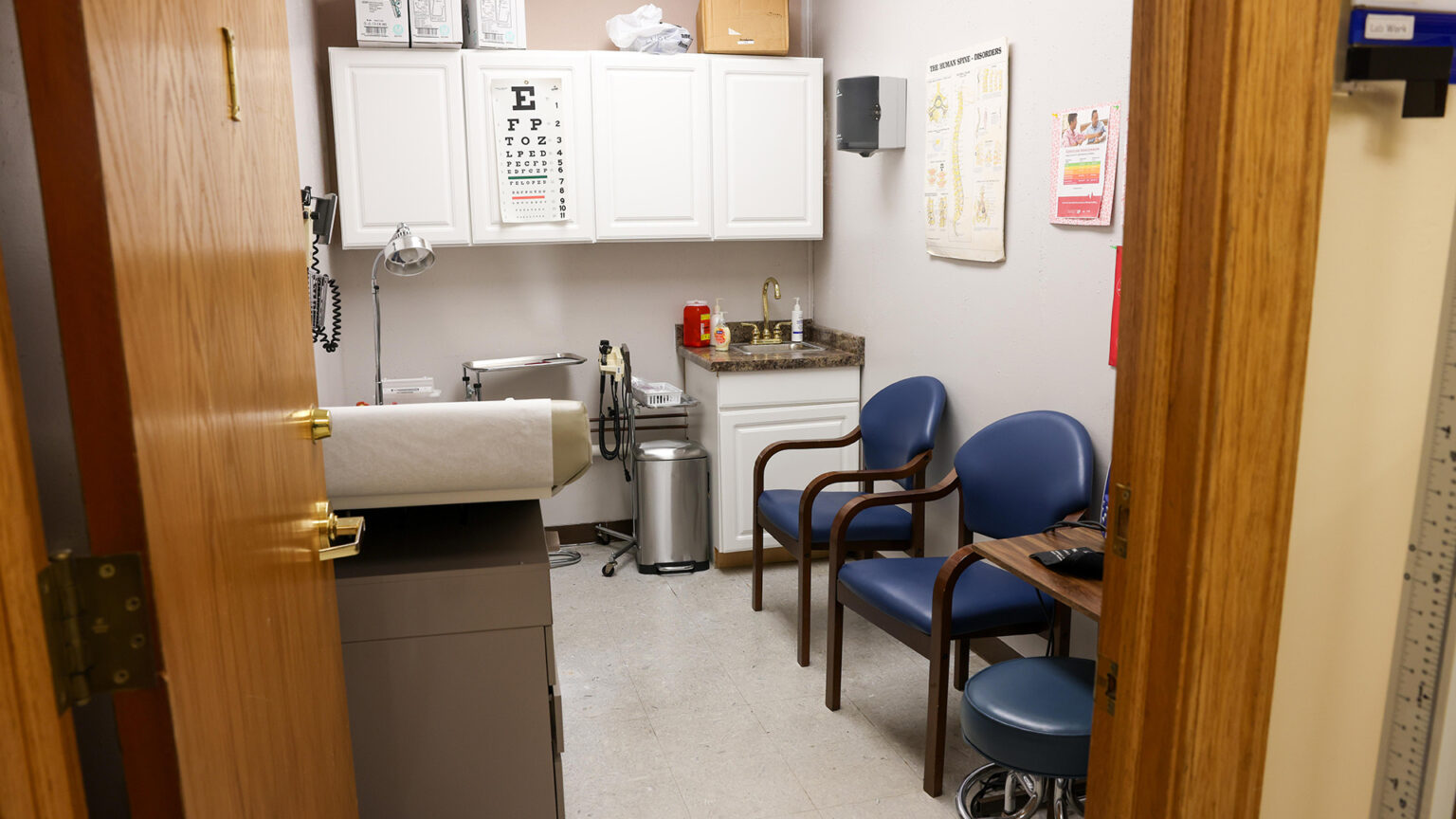 A room with an exam table, two wood arm chairs with padded seats and backs, and a padded stool under a table sit in a room with cabinets, a sink, and other items along a back wall, with an eye chart displayed on a cabinet door and a graphic showing a spinal column on a wall, with an open wood door and its frame in the foreground.