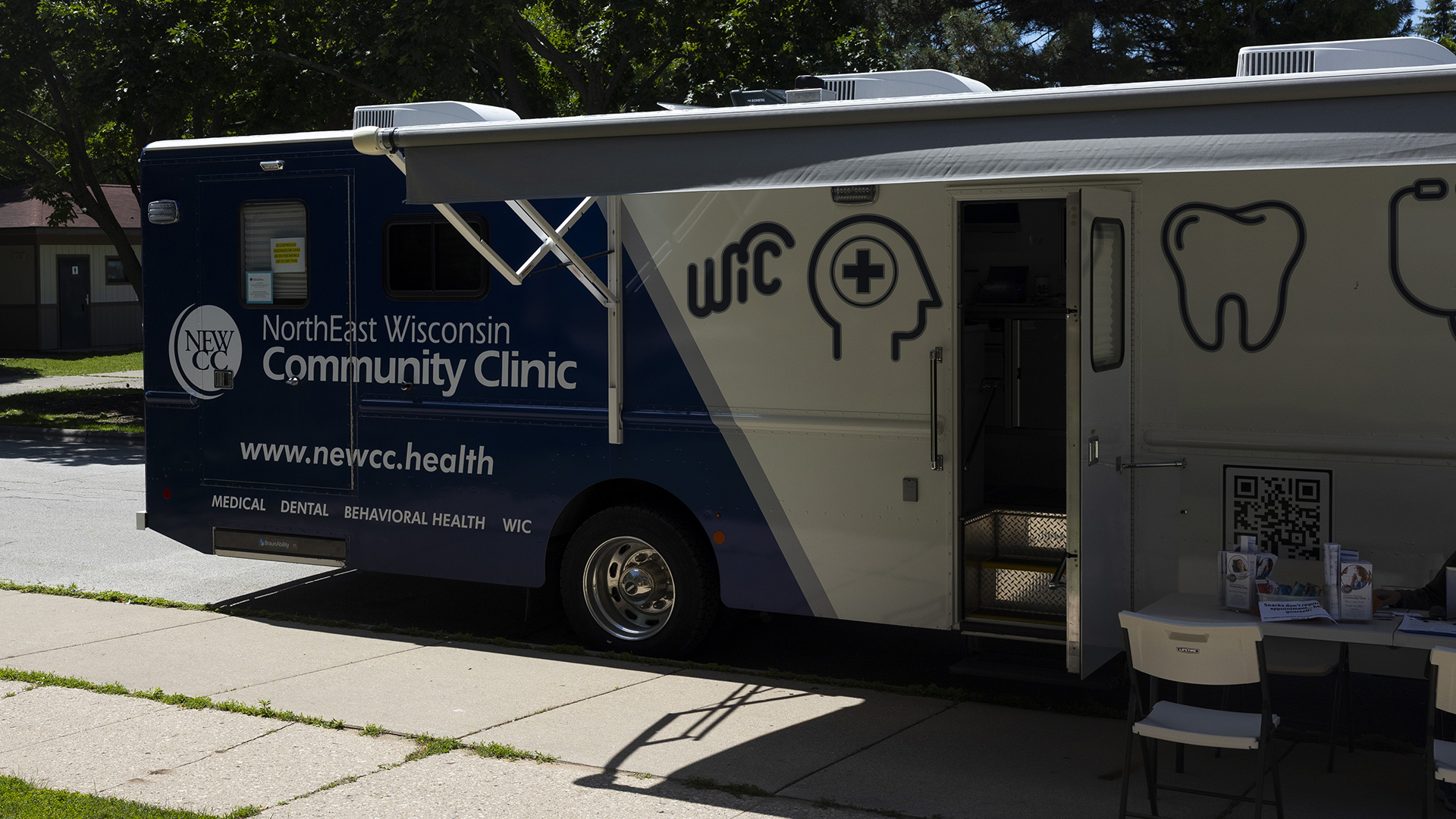A recreational vehicle with the words "NorthEast Wisconsin Community Clinic," the URL www.newcc.health and different graphics is parked on the side of a road next to a sidewalk, with its open side awning providing shade to a folding table and chairs in place next to and open door, with trees and a building in the background.