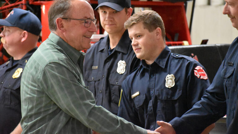 Tom Tiffany shakes the hand of a firefighter wearing a station uniform, with three other firefighters standing in the background in front of an out-of-focus vehicle.