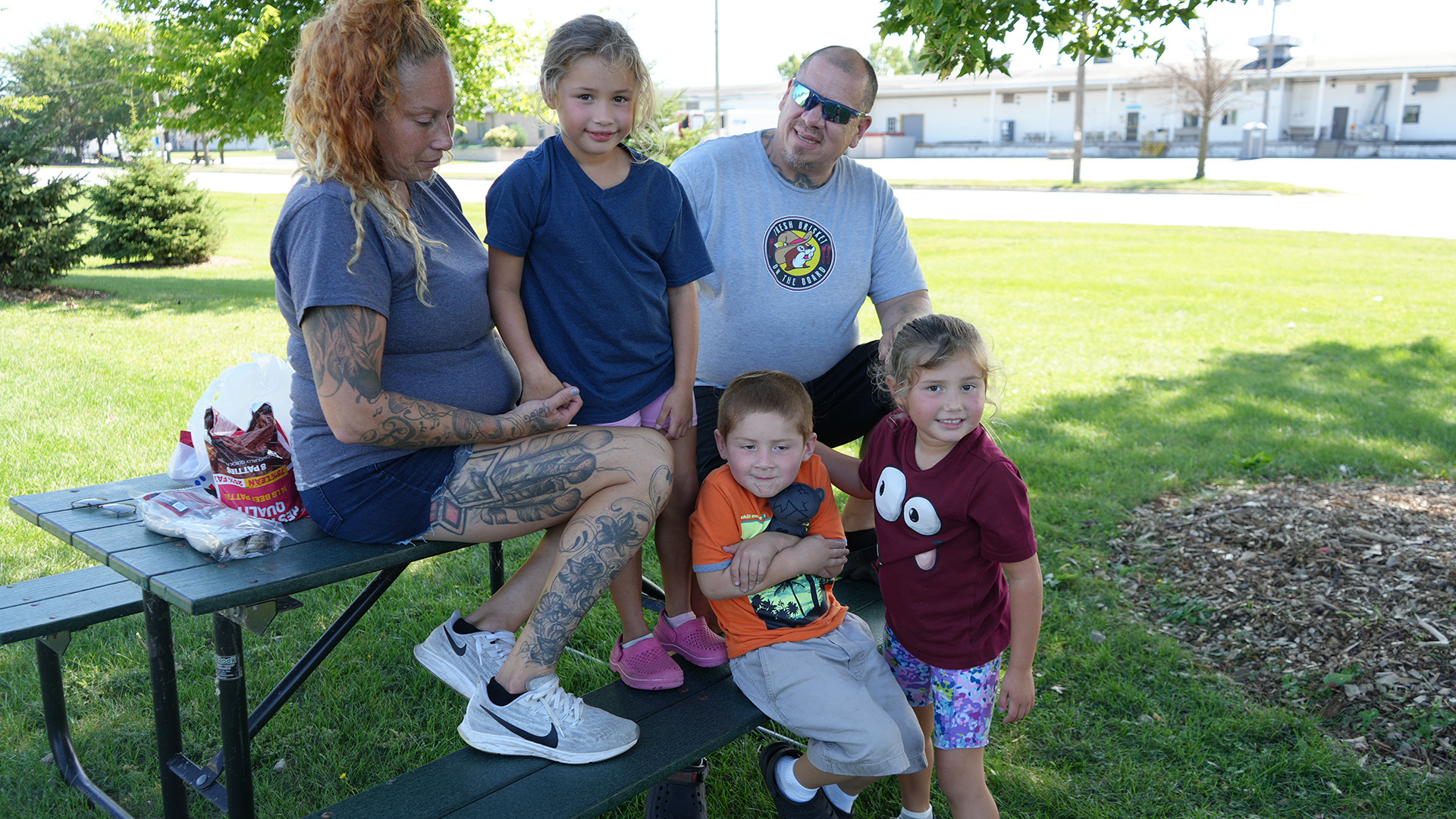 Estelle Moncada, James Gray and three children pose for a portrait while seated on and standing next to a wood and metal picnic tale on a lawn next to a mulch bed and under overhanging leaves, with short conifer trees behind them and a sunlit road and long two-story building in the background.