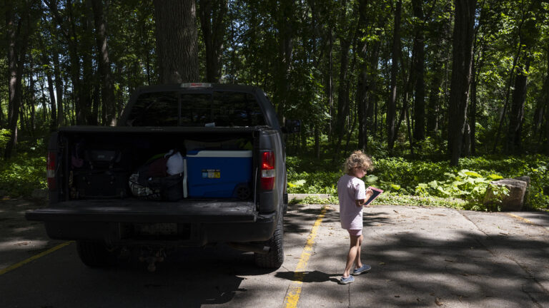 A child walks in a leaf-shaded asphalt parking lot next to a pickup truck with an open rear gate, with a wooded area in the background.