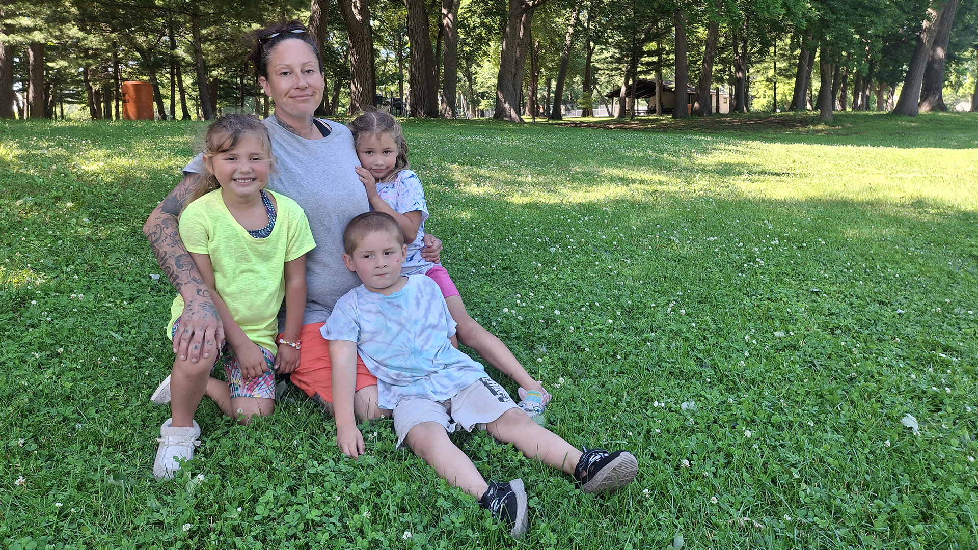Estelle Moncada and three children pose for a portrait while seated on a lawn, with a wooded area in the background.