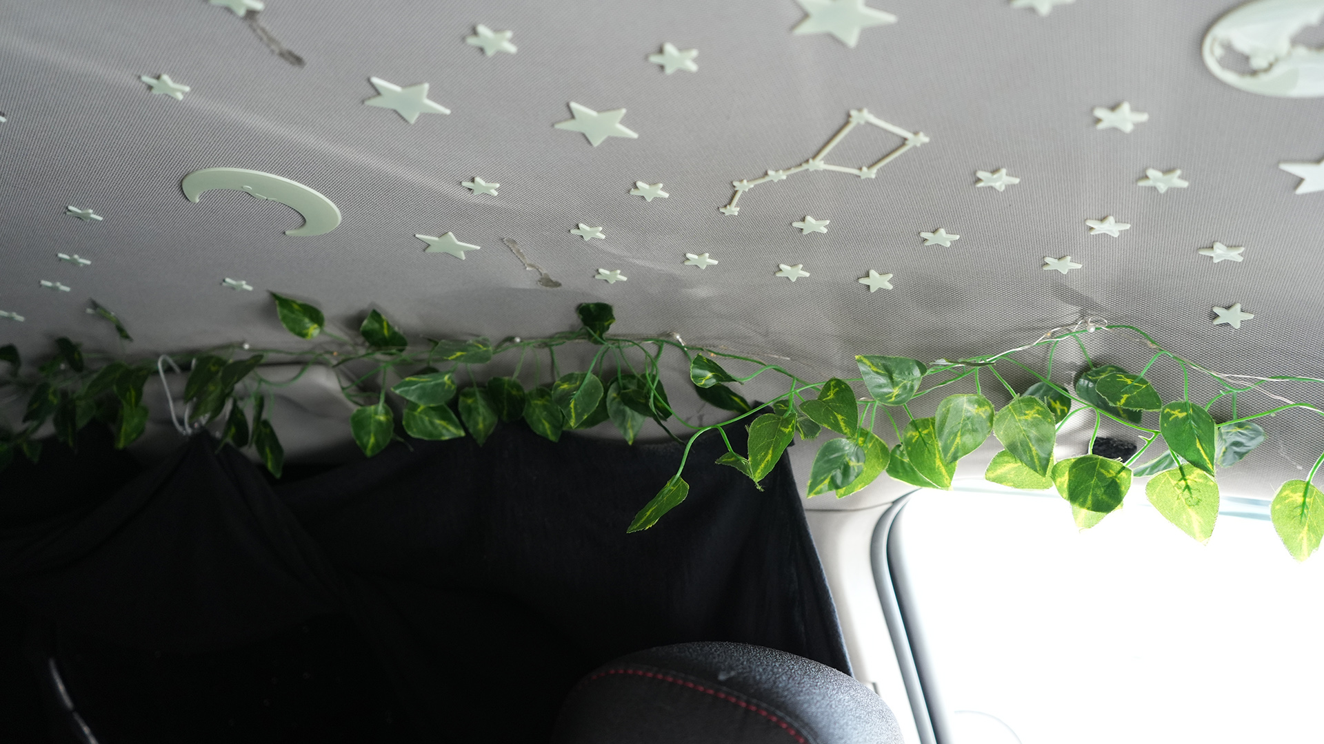Glow-in-the-dark decals of stars, Ursa Major, the moon and Earth are affixed to the interior roof of a vehicle next to a garland of faux foliage above a window and car headrest.