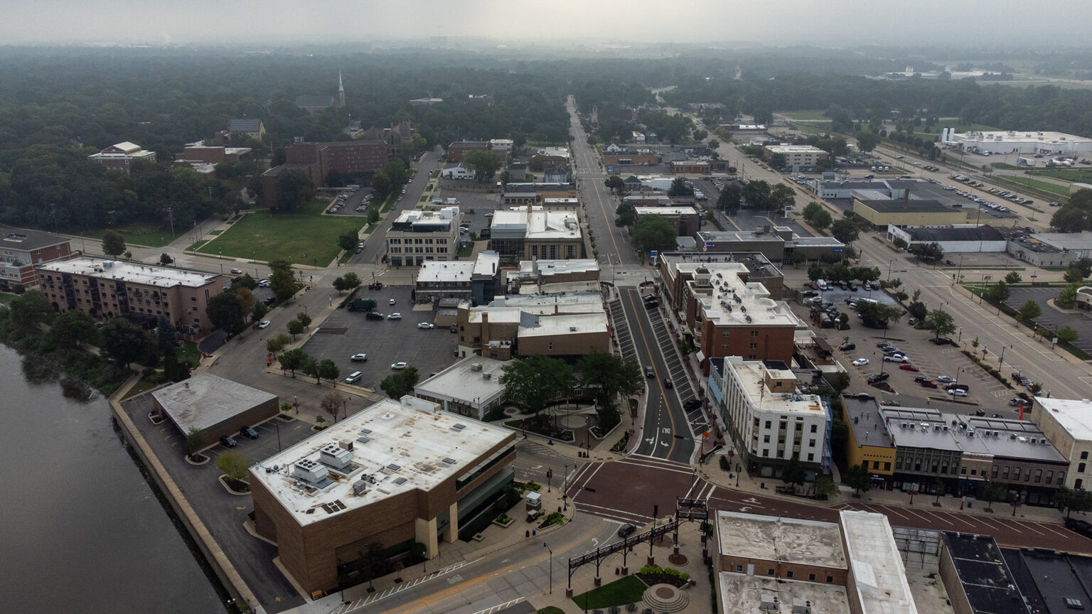 An aerial photo shows a cluster of multi-story buildings along multiple blocks of a grid of streets next to a body of water, with tree-covered neighborhoods extending to the horizon under a hazy sky. An aerial photo shows a cluster of multi-story buildings along multiple blocks of a grid of streets next to a body of water, with tree-covered neighborhoods extending to the horizon under a hazy sky.