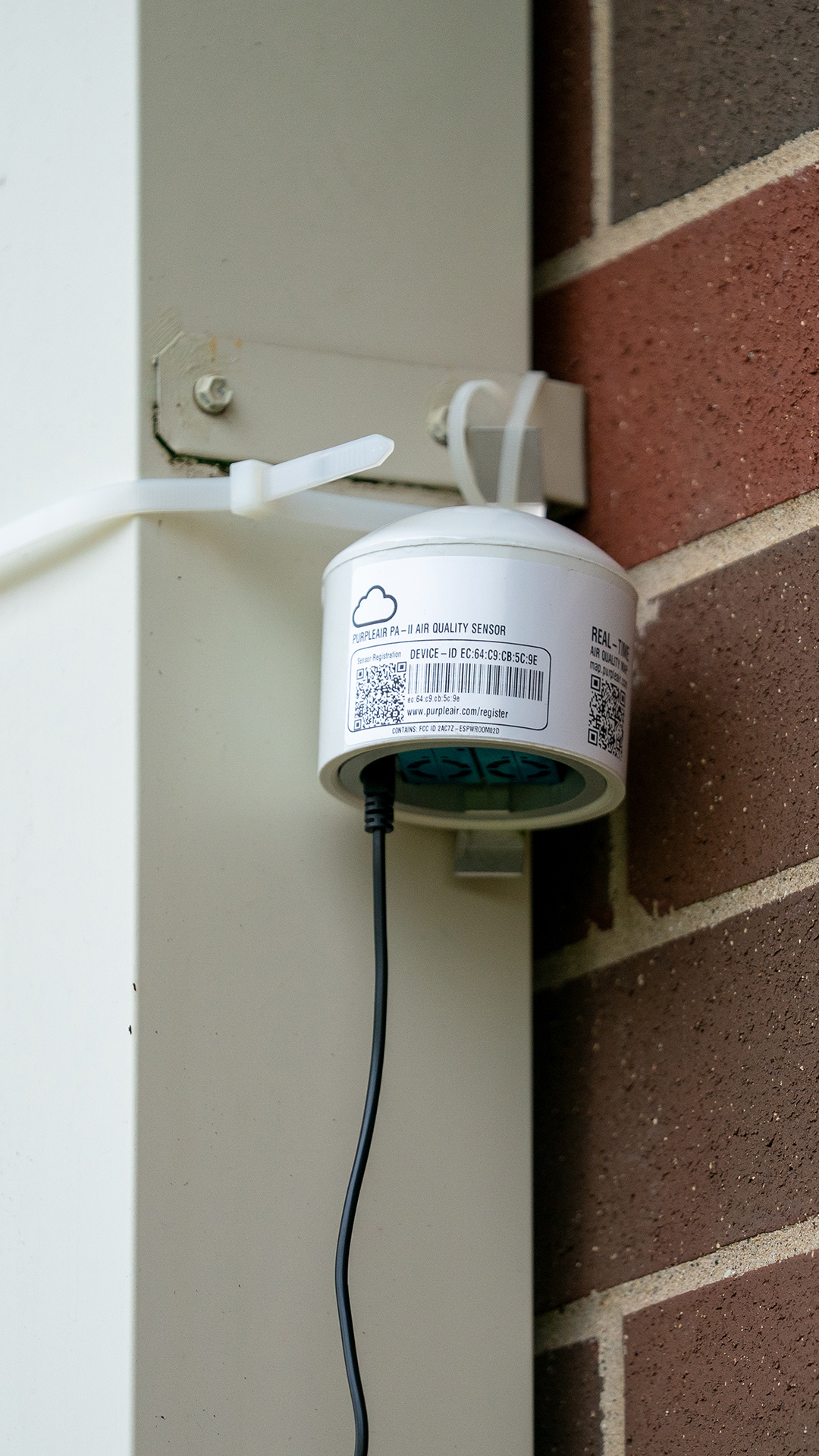 A cylindrical metal object with a low conical top and a power cord extending from its bottom is connected with zip ties to a metal brace mounted on a brick wall.