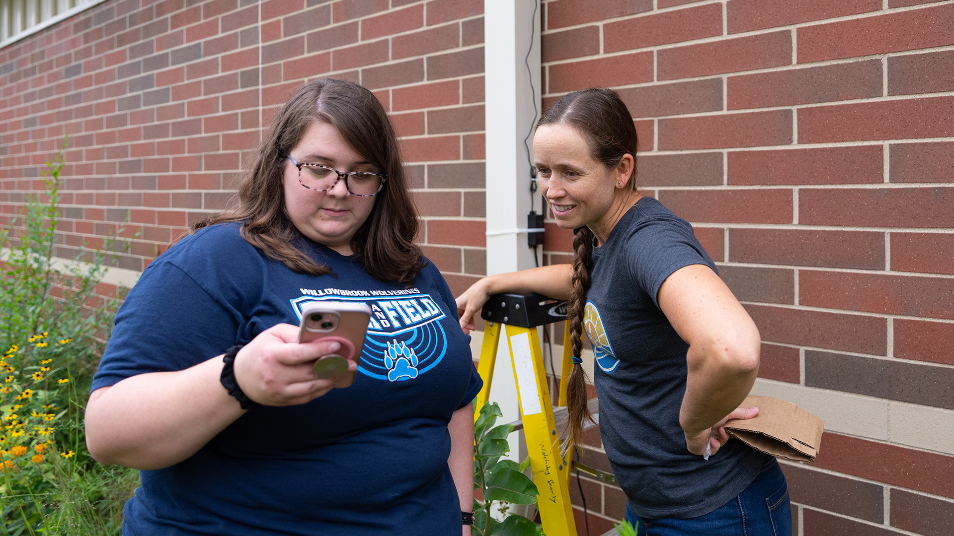 Erika Goodsell looks at the screen of a smart phone in her right hand while standing next to Brittany Keyes, who leans on the top of a step ladder with her right forearm, with each standing among wild grasses and flowers in front of a brick wall.