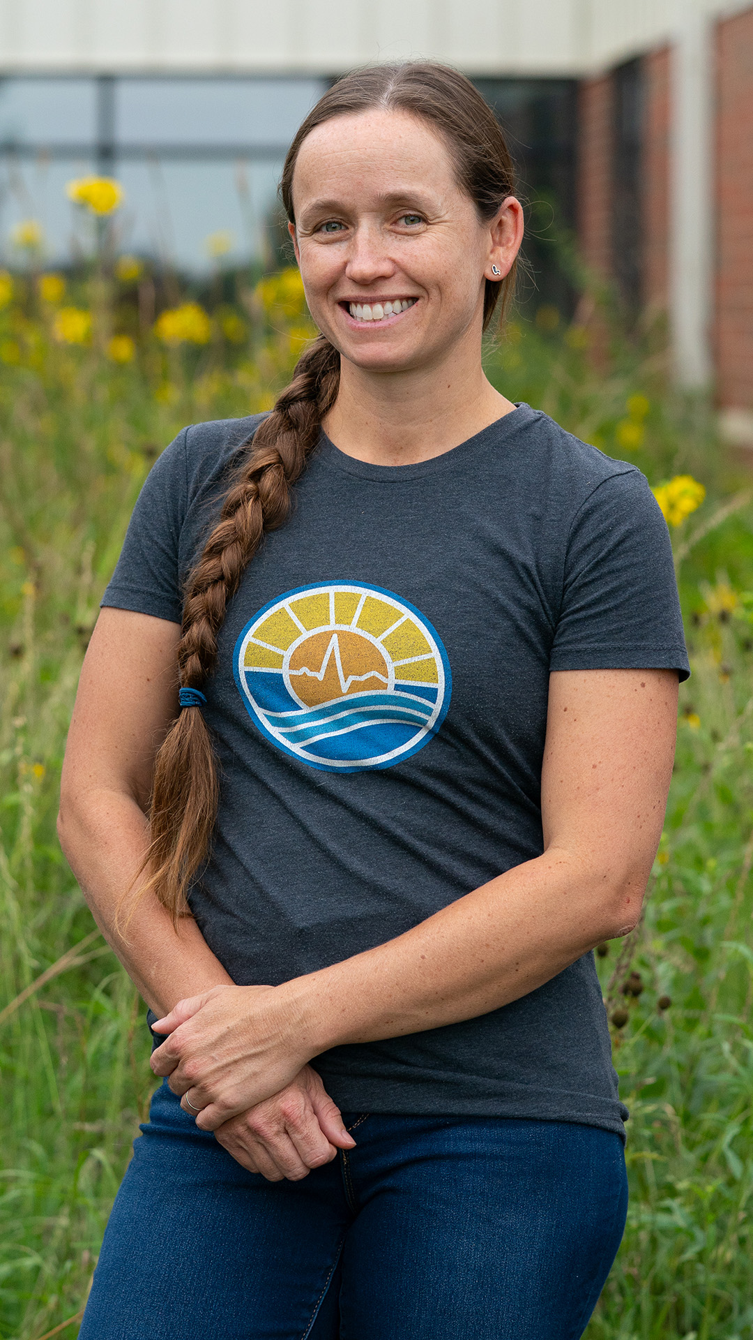 Brittany Keyes poses for a portrait while standing in front of an out-of-focus garden of native grasses, with an out-of-focus brick and glass building wall in the background.