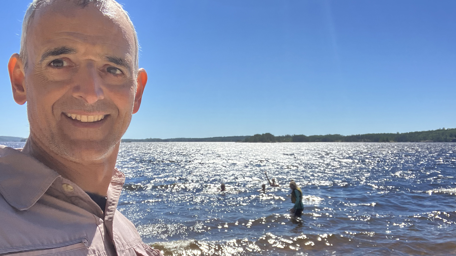 Pablo Toral smiles in a selfie pose in front of a sunlit body of water with multiple people standing and swimming in low waves, with wooded hills on the far shoreline.