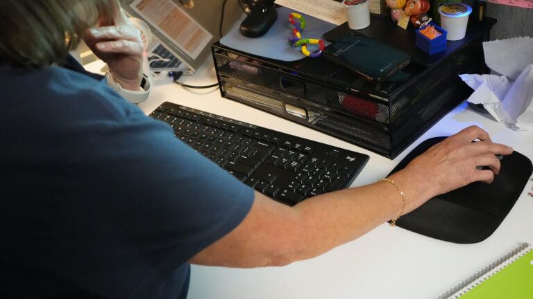 The right arm of a person holds a wireless computer mouse on a mouse pad while sitting and facing a desk, with a wireless keyboard, metal desktop storage trays and other items on its surface.
