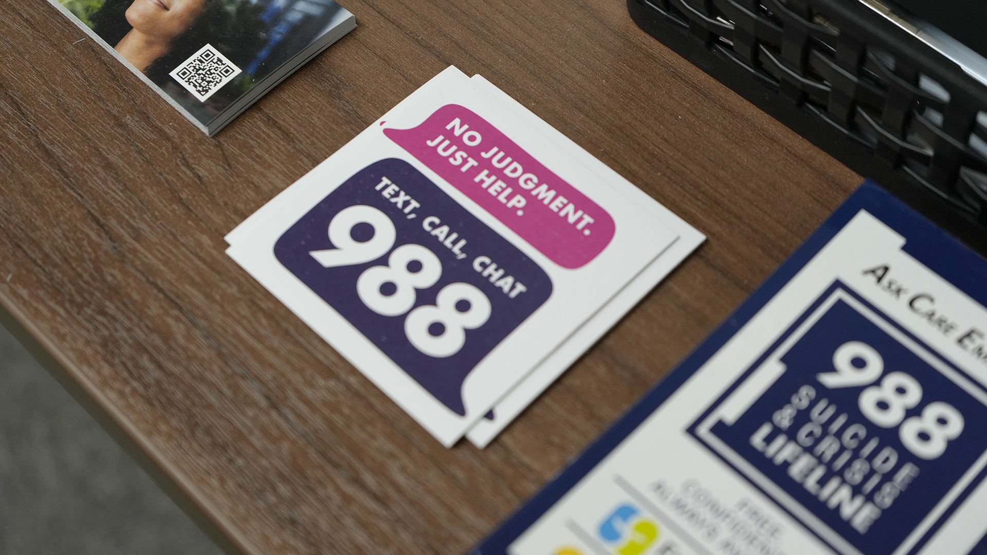 Square cards printed with the words "No Judgement. Just Help." and "Text, Call, Chat 988" inside talk bubbles sit in a short stack on a laminated wood desk service next to stacks of other out-of-focus printed cards.