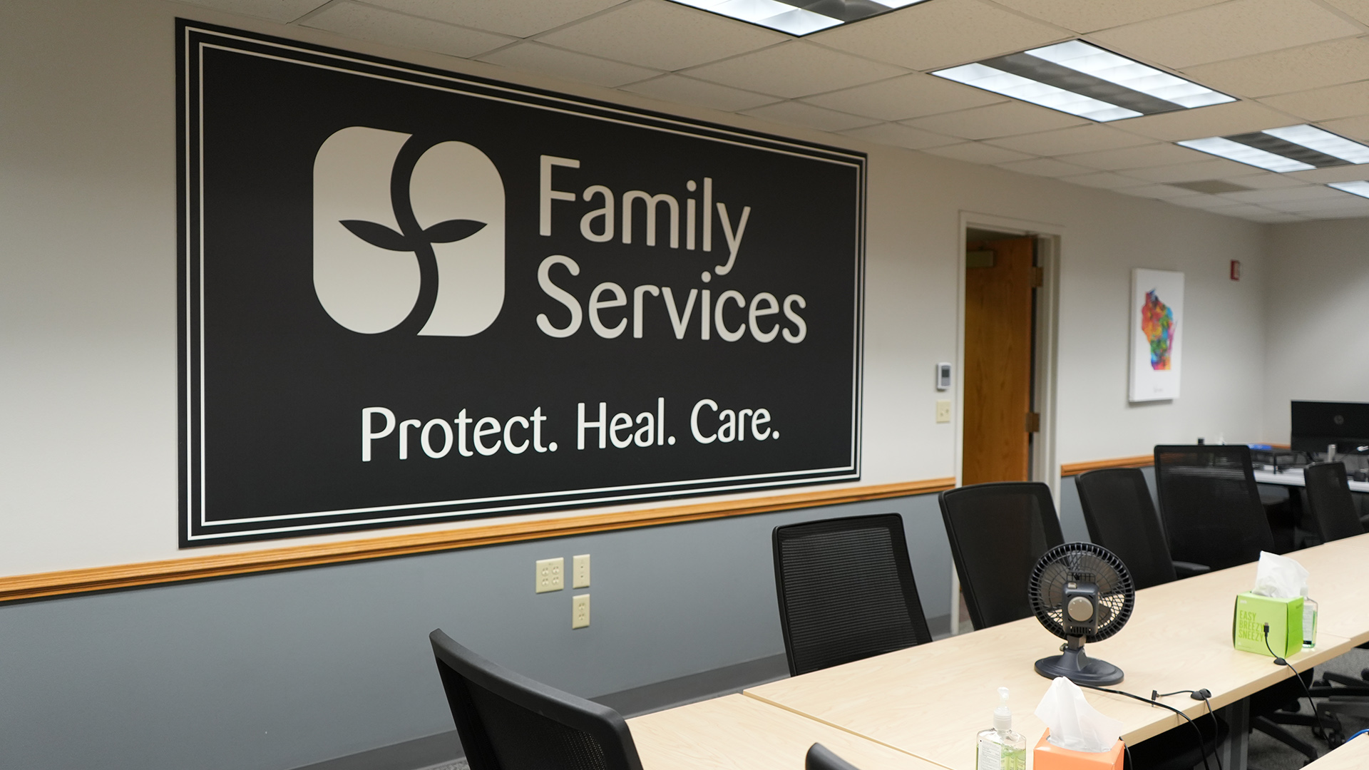 A large sign with the words "Family Services" and "Protect. Heal. Care." alongside a stylized logo of an "F" in the shape of a plant shoot is mounted on a wall in a room with a conference table surrounded by high-backed rolling chairs, a canvas graphic with the outline of Wisconsin mounted on a wall next to an open wood door, and a drop-panel ceiling with fluorescent lights.