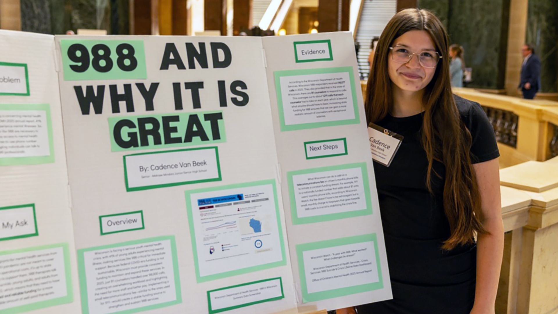 Cadence Van Beek stands next to a research poster with the title "988 and Why It Is Great" in front of a marble balustrade in a room with marble masonry walls and out-of-focus people standing in the background.