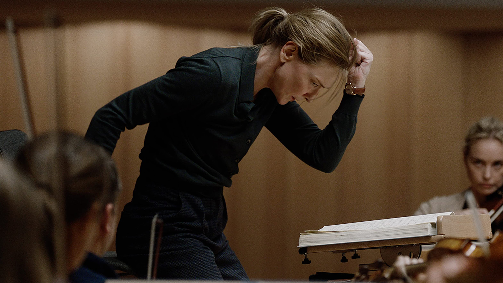 A conductor in black leans intensely over a score on a podium, fist raised to her head, as orchestra musicians with string instruments watch nearby.