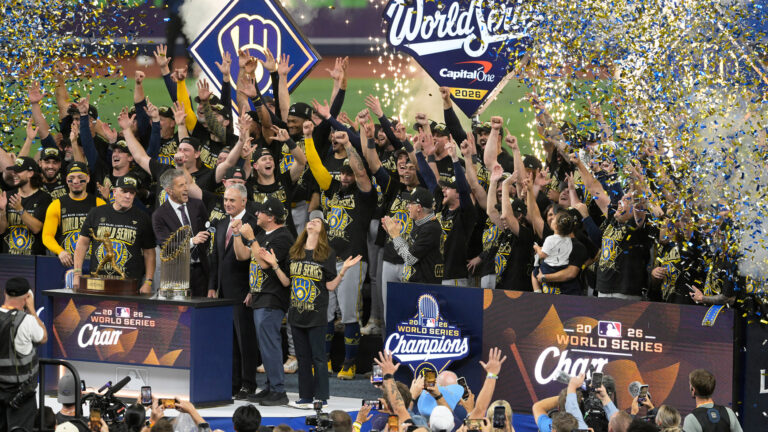Milwaukee Brewers owners, staff, players and their families, all wearing World Series champion shirts, celebrate on a stage behind a dais with the Commissioner's Trophy and Willie Mays Most Valuable Player Award Trophy on its surface, with the team's ball-in-glove logo and World Series logos illuminated on signs in the background with blue and gold confetti and fireworks sparklers flying in the air, with photographers and people cheering facing the group in the foreground.