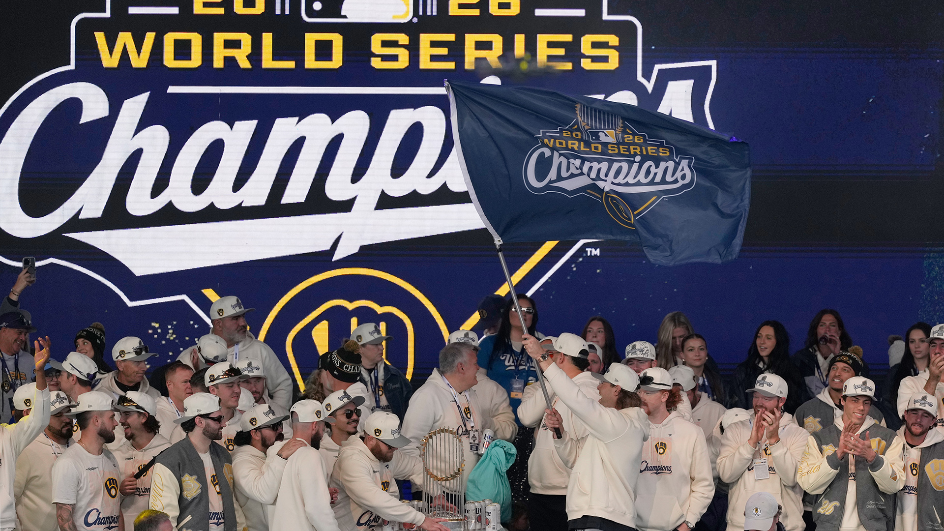 Trevor Megill holds a metal pole and waves a flag with a logo that shows the Major League Baseball Commissioner's Trophy and the Milwaukee Brewers and MLB logos alongside the words "2026 World Series Champions," with numerous cheering and applauding people standing next to and behind him, with the Commissioner's Trophy standing on a table in the midst of the group, and with the same champions logo displayed on a screen in the background.
