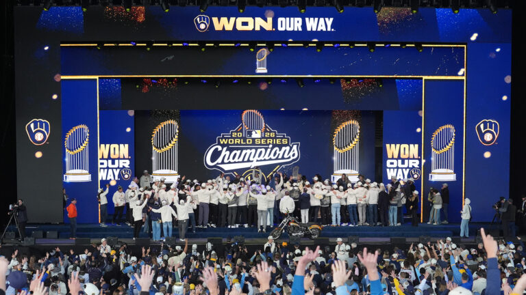 People standing on a stage and a larger number of people standing in an audience raise their hands in the air while facing a stage backdrop projecting images of the baseball glove logo of the Milwaukee Brewers, the Major League Baseball Commissioner's Trophy and a logo showing both along with the words 2026 World Series Champions alongside the words Won. Our Way.
