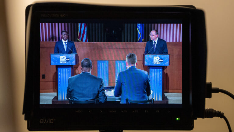 A video camera monitor with two cables connected to one side and an illuminated power light shows David Crowley and Tom Tiffany standing behind podiums with two microphones affixed to the top of each and the logo on WISN 12 ABC on their front in a room with a wood judicial dais and the U.S. and Wisconsin flags in the background, with two people seated in chairs facing them in the foreground.