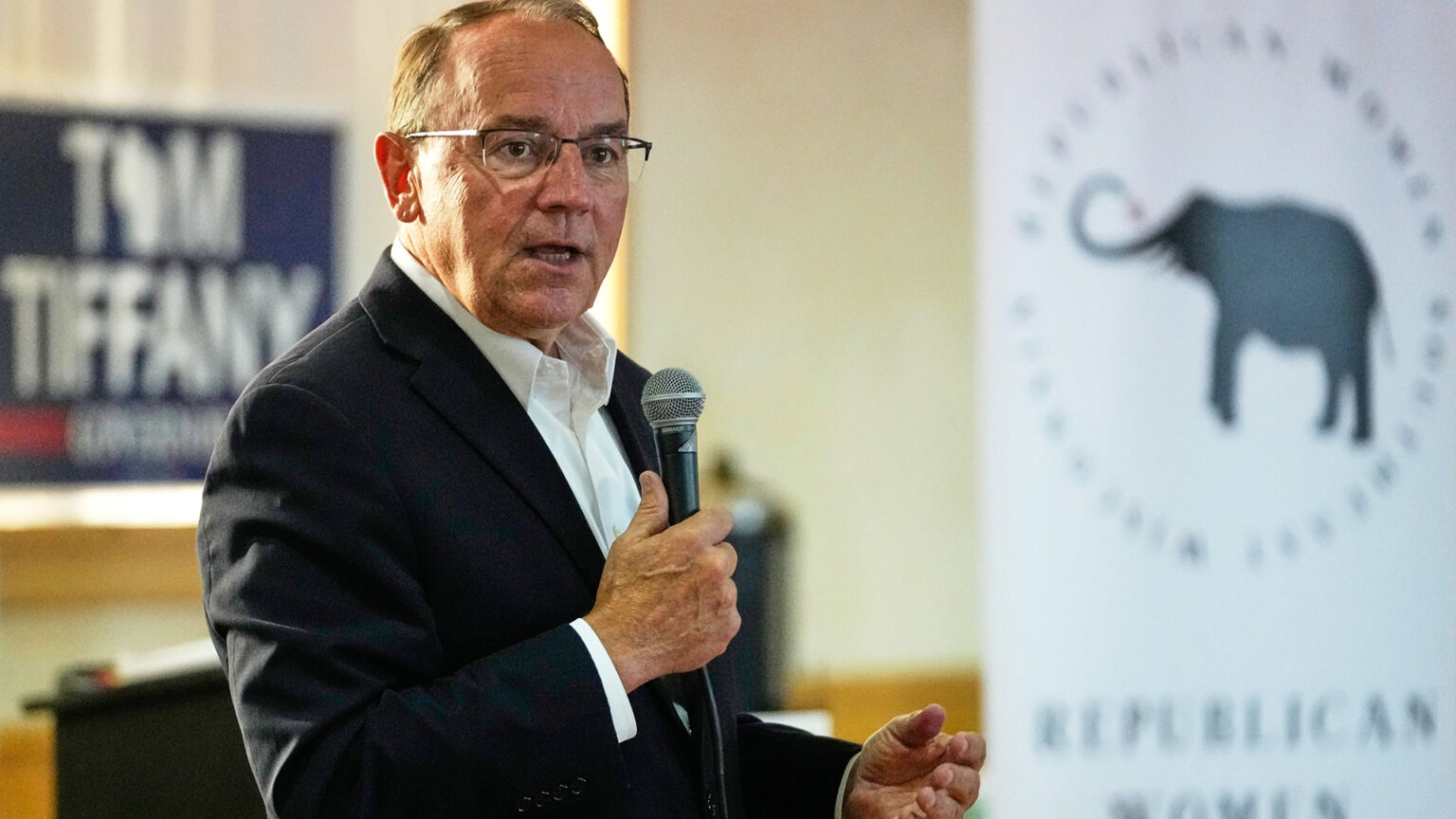 Tom Tiffany gestures with his left hand and speaks into a wired microphone held in his right hand, while standing in a room with an out-of-focus campaign yard sign and Republican Party banner with a graphic of an elephant displayed in the background.