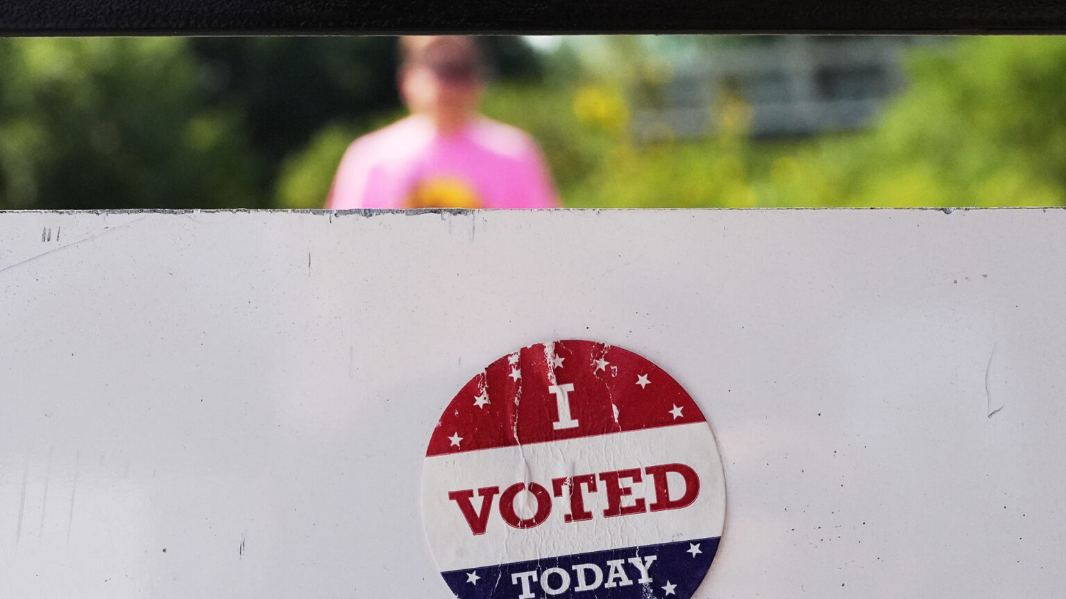 A scuffed sticker with the words I Voted Today is attached to the back of sign, with an out-of-focus person visible outside beneath the bottom edge of the upper edge of a window.