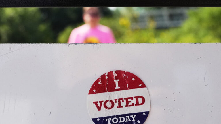A scuffed sticker with the words I Voted Today is attached to the back of sign, with an out-of-focus person visible outside beneath the bottom edge of the upper edge of a window. A scuffed sticker with the words I Voted Today is attached to the back of sign, with an out-of-focus person visible outside beneath the bottom edge of the upper edge of a window.