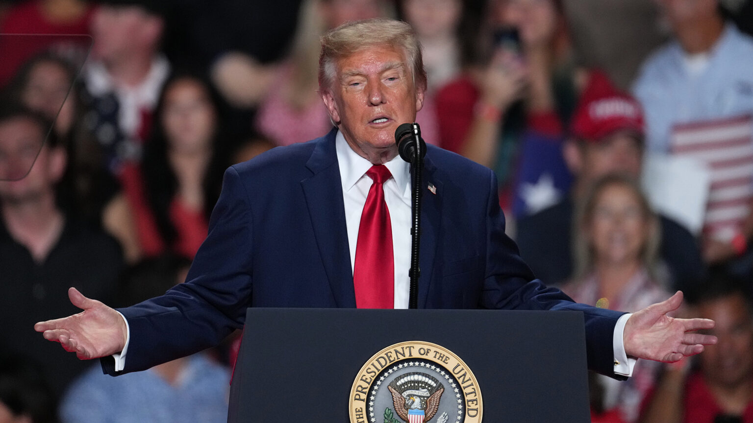 Donald Trump gestures with both hands held out to the side while speaking into a microphone mounted to the top of a podium with the Seal of the President of the United States affixed to its front, with out-of-focus audience members in the background behind him and a teleprompter mirror in the foreground on one side.