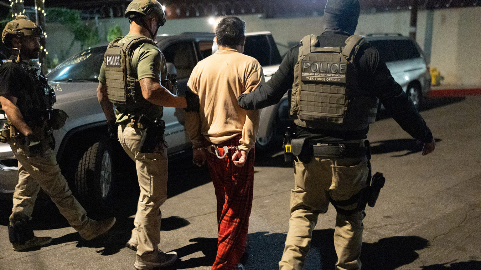 Two law enforcement officers wearing tactical vests and utility belts hold both arms of a person whose hands are restrained in two pairs of handcuffs behind their back, with another officer standing to the side, with parked vehicles and an out-of-focus concrete wall topped with razor wire in the background.