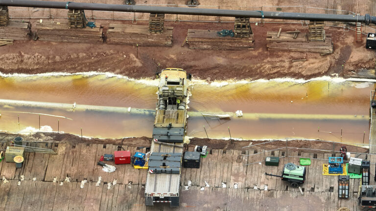 An aerial photo shows a the front half of a flatbed trick spanning a course of liquid filling a trench over a below-grade pipeline at a construction site with planks of wood serving as a corduroy road and stacks of wood planks next to a section of an above-grade pipeline siting on stacks of wood pallets. An aerial photo shows a the front half of a flatbed trick spanning a course of liquid filling a trench over a below-grade pipeline at a construction site with planks of wood serving as a corduroy road and stacks of wood planks next to a section of an above-grade pipeline siting on stacks of wood pallets.
