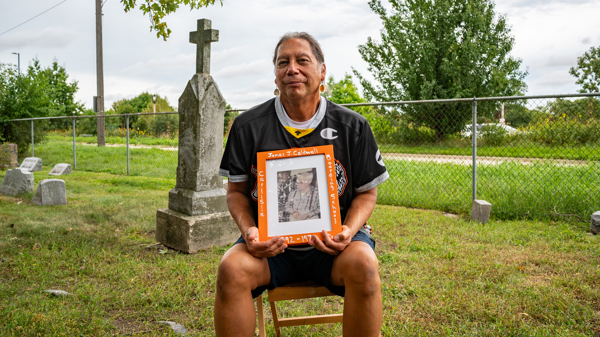 Mark Denning holds a photo in his arms while sitting in a folding chair in a cemetery, with a stone obelisk monument topped by a cross and other headstones behind him, with a chain-link fence, road and trees in the background.
