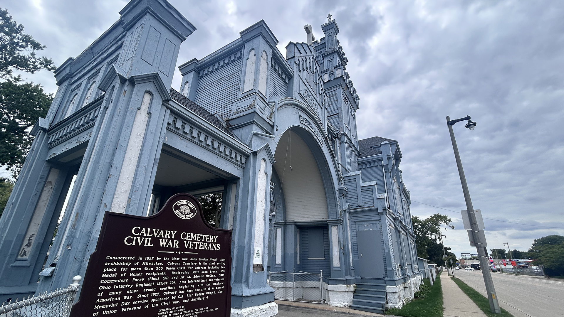 A multi-story painted wood-sided building with crenelated and non-crenelated towers and an archway over a paved entrance by a road, with trees and buildings in the background and a historical marker in the foreground with the title text "Calvary Cemetery" and "Civil War Veterans" followed by additional text.