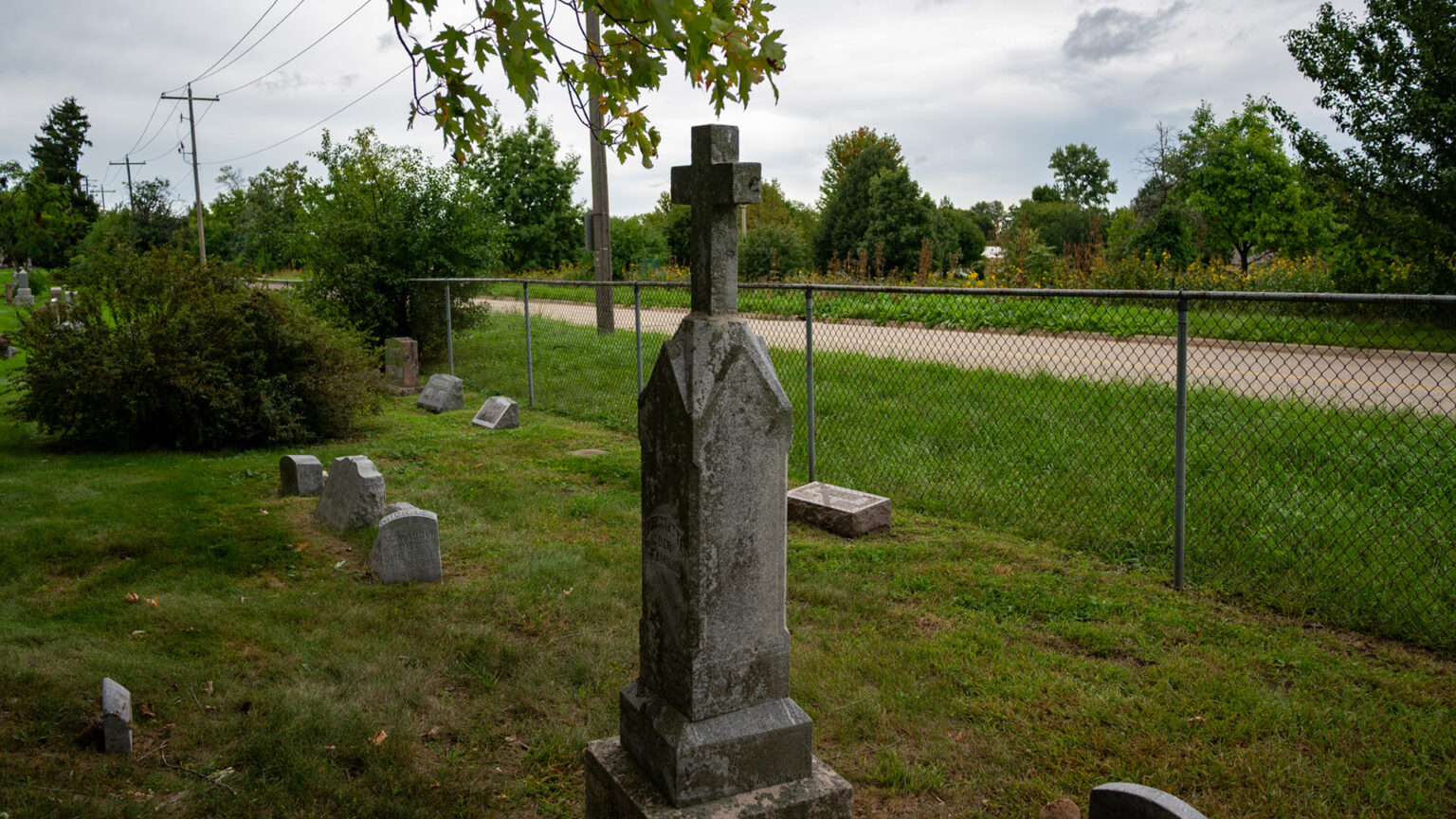 A stone obelisk monument topped by a cross stands among lower headstones in a cemetery next to a chain-link fence, with a line of utility poles, road and trees in the background. A stone obelisk monument topped by a cross stands among lower headstones in a cemetery next to a chain-link fence, with a line of utility poles, road and trees in the background.