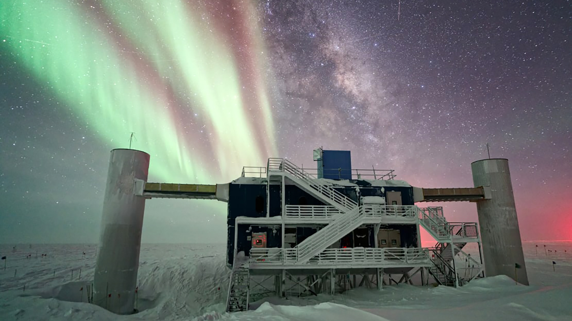 Bands of aurora australis shimmer next to the Milky Way band of stars in a night sky over a building with two concrete pillars on either side of a superstructure with exterior stars and walkways encased in an ice landscape.