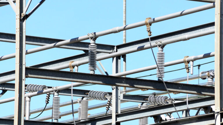 Light and shadow provide contrast to a superstructure of metal girders, poles and coils that connect in an irregular grid, with a clear sky in the background.