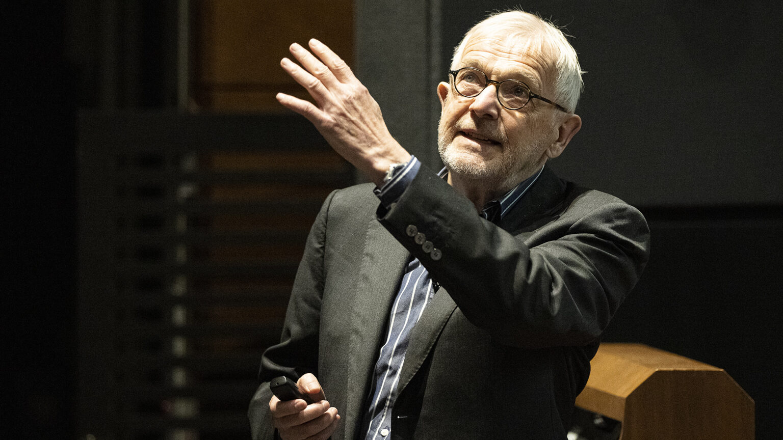 Francis Halzen stands and gestures with this left hand and holds a presentation clicker in his right hand while speaking in a room with an out-of-focus wood podium in the background. Francis Halzen stands and gestures with this left hand and holds a presentation clicker in his right hand while speaking in a room with an out-of-focus wood podium in the background.