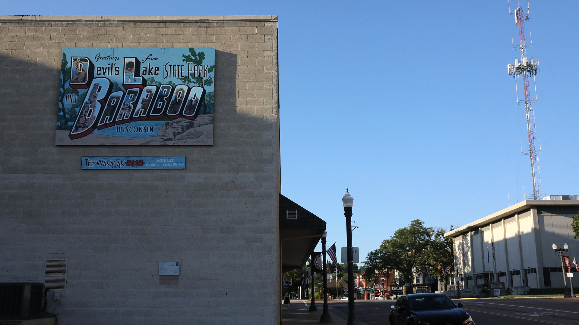 A vintage bubble letter postcard-style promotional sign with the words "Greetings from Devil's Lake State Park," "Baraboo" and "Wisconsin" is mounted to the concrete block wall of a two story building, with a car parked in front of it on an adjacent street next to a street lamp, with trees, other buildings, and a multi-story building across and down the street with a cell tower rising behind it.