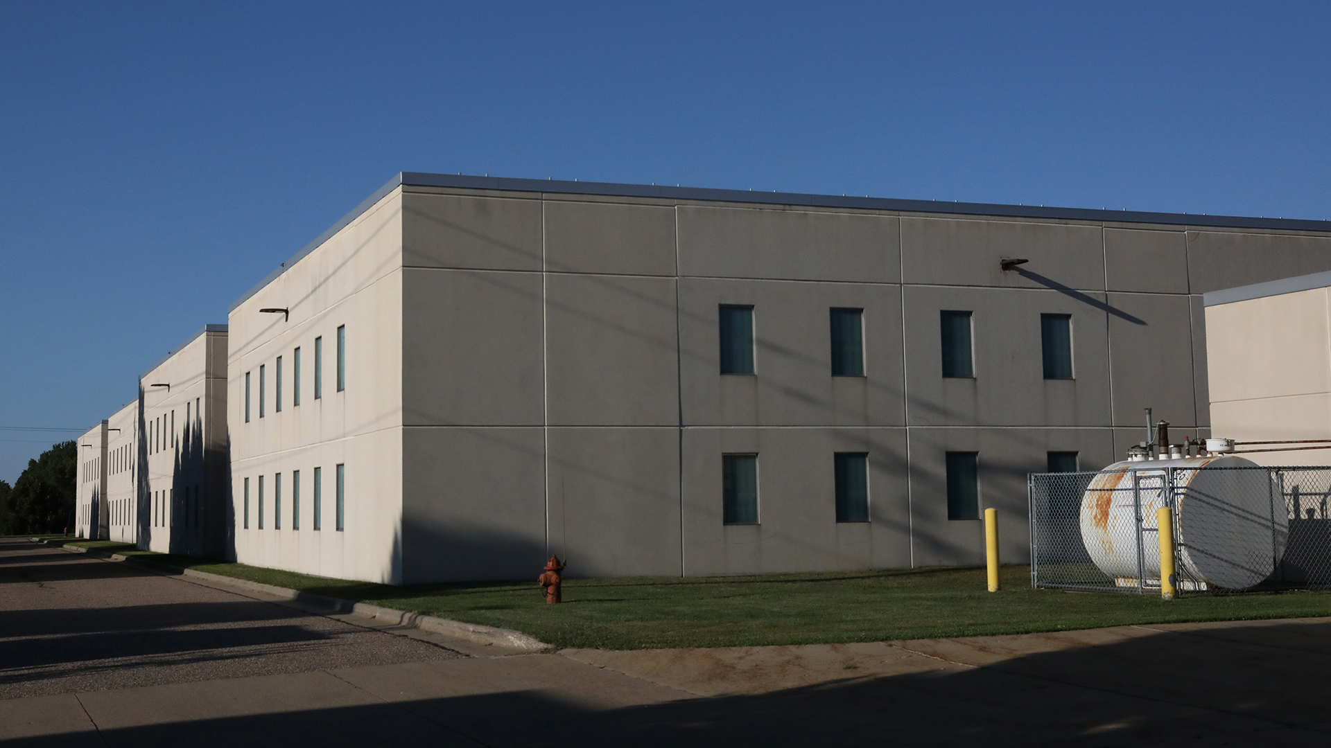 A two-story stone-sided building with rows of windows on multiple walls stands next to a road, with storage tank for compressed gas surrounded by a chain-link fence on a lawn in the foreground.
